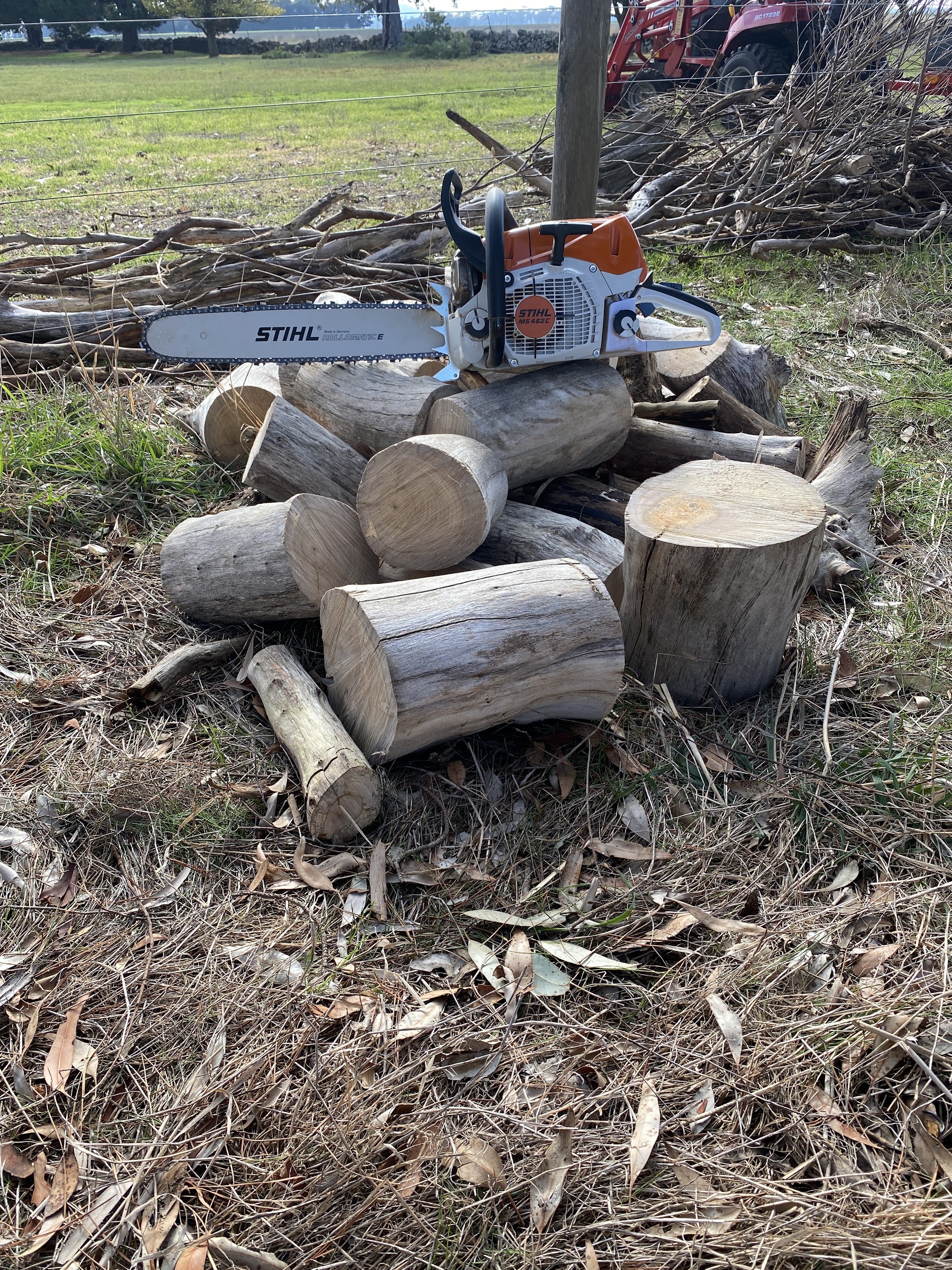 The same Stihl chainsaw on a pile of cut firewood rounds, with a paddock and a tractor behind