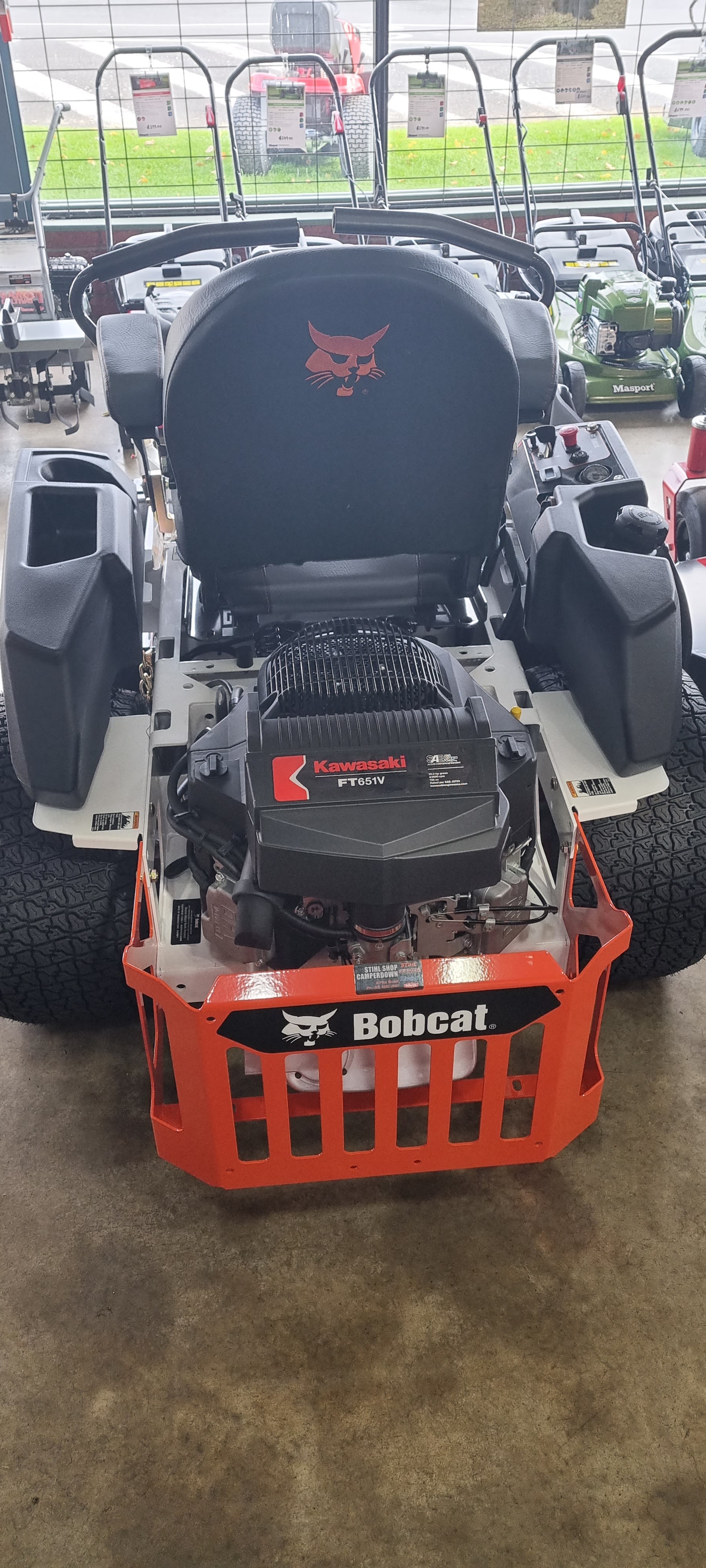 A Bobcat zero turn mower head on, showing its Kawasaki engine, with green Masport push mowers behind it